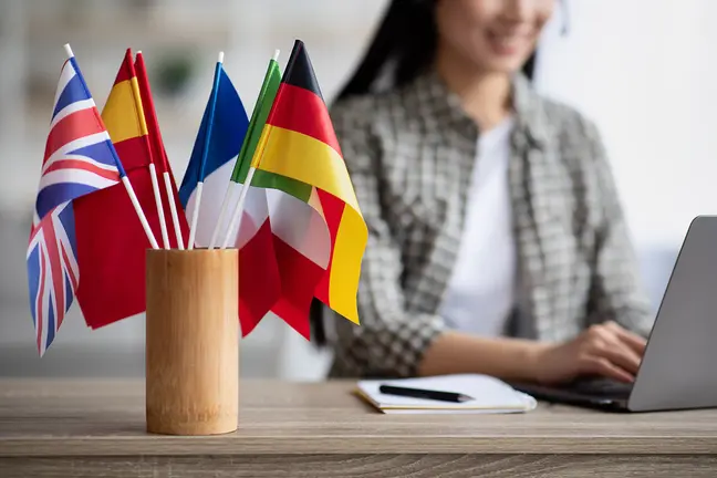 Cropped of asian young woman foreign language teacher sitting at workdesk and using laptop, selective focus on international flags. Foreign language online course, class, e-education concept