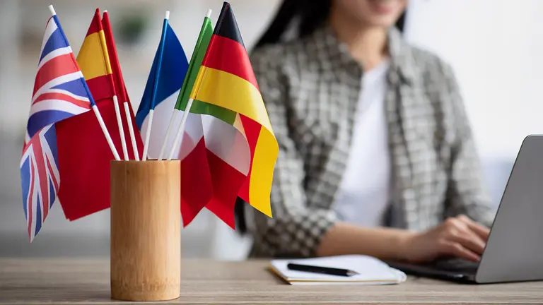 Cropped of asian young woman foreign language teacher sitting at workdesk and using laptop, selective focus on international flags. Foreign language online course, class, e-education concept